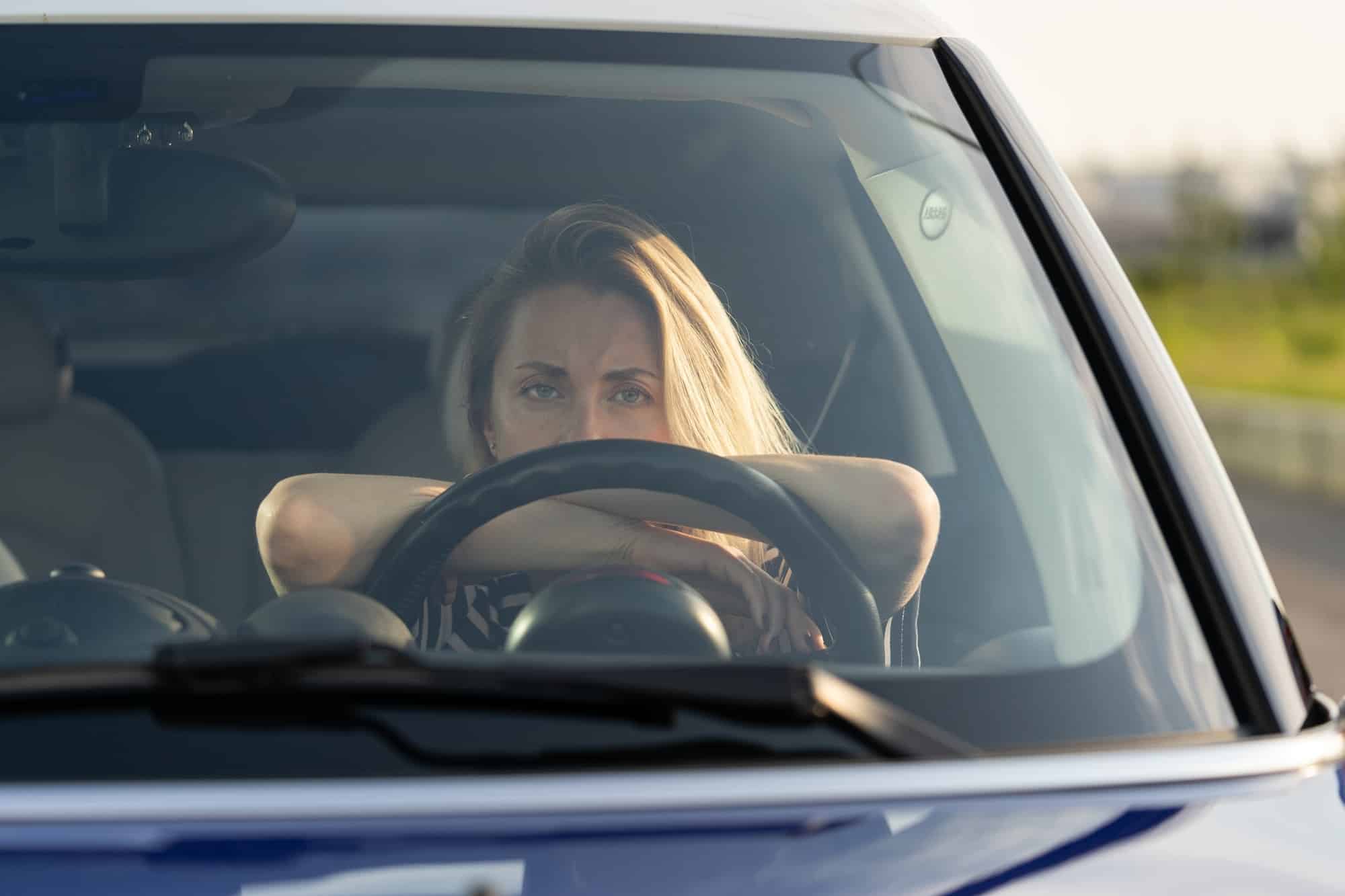 Thoughtful tired woman driving car, looking through windshield at camera, thinking about something.